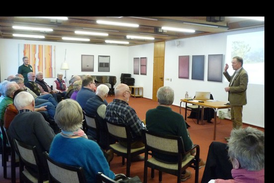 Ein Referent steht in einem Raum vor einer Leinwand. Vor ihm sitzen Zuhörer auf Stühlen.